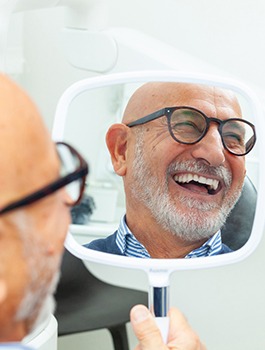 Bald man in glasses smiling at reflection in handheld mirror at dental office